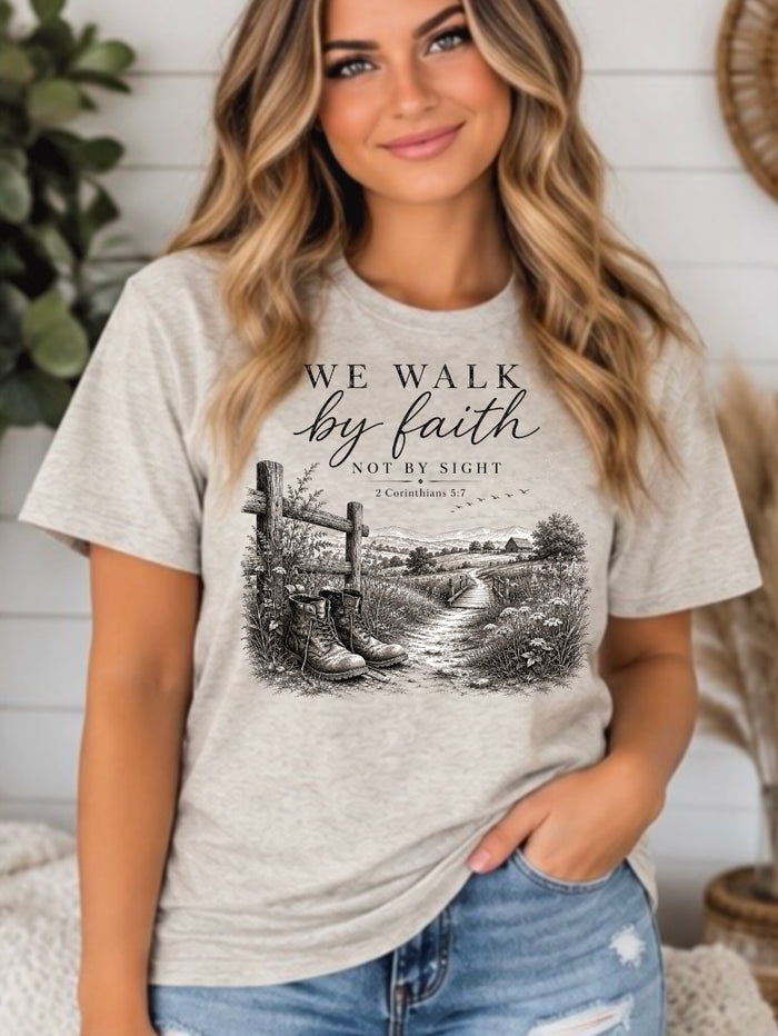 A woman wearing a light heather oatmeal graphic t-shirt with a black and white print of boots at a crossroads and the text 'WE WALK by faith NOT BY SIGHT'.