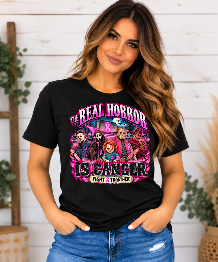 A woman wearing a black graphic t-shirt with a pink and multicolor design that reads 'THE REAL HORROR IS CANCER FIGHT TOGETHER' with illustrated horror characters, paired with blue jeans, set against a white wood-paneled wall background.